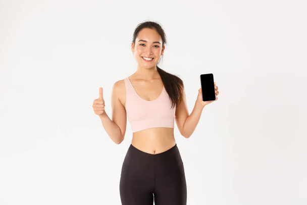 Sport, Technologie und aktives Lifestylekonzept. Zufrieden lächelndes asiatisches Fitness-Girl, attraktive Sportlerin mit Daumen hoch und Smartphone-Bildschirm, empfehle mobile Workout-Tracker-App - Foto, Bild