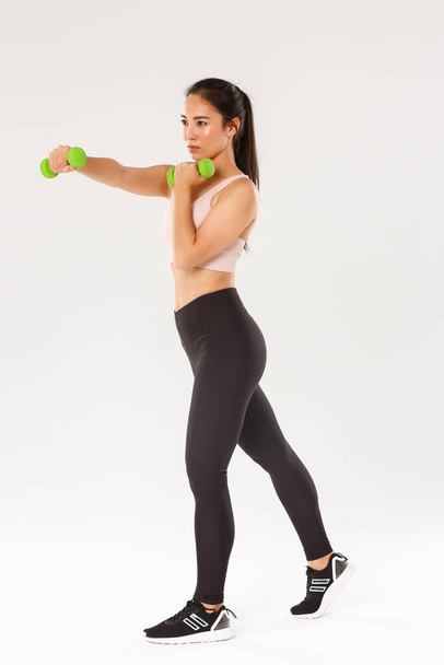 Sport, gym and healthy body concept. Full length of focused female athelte, asian brunette girl doing fitness exercises, workout with dumbbells, productive gym training over white background - Photo, Image