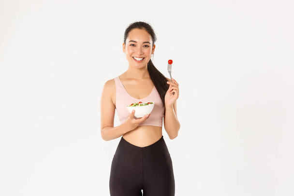 Sport, wellbeing and active lifestyle concept. Smiling healthy and slim asian girl in fitness clothing, holding salad and pick tomato on fork, staying fit with diet and special workout - Photo, Image
