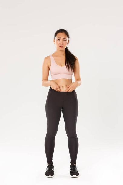 Full length of upset and gloomy asian girl in fitness clothing, showing fat on belly, complaining on body, frowning and looking disappointed, starting workout, trying lose weight, white background - Photo, Image