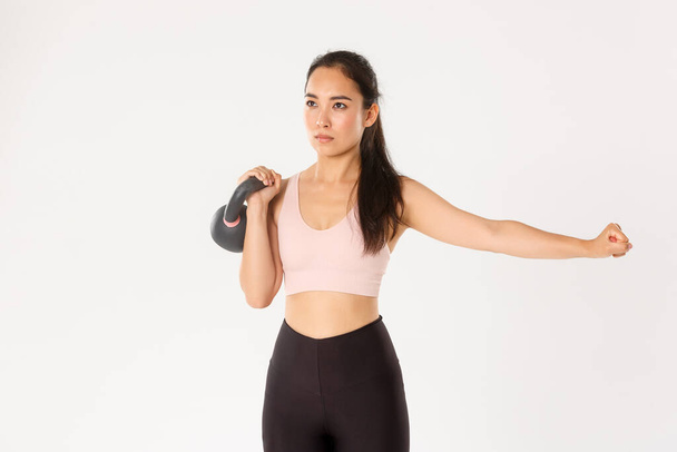 Sport, wellbeing and active lifestyle concept. Focused and motivated asian female workout with kettlebell, lift weight and extend one hand, repeat exercise after coach in gym, white background - Foto, Imagen