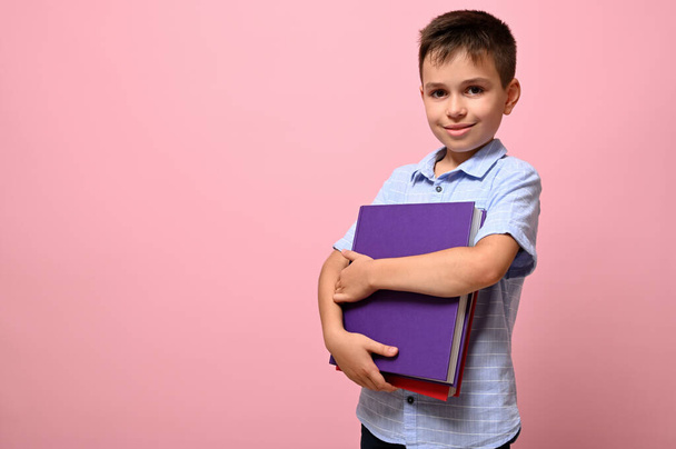 An adorable schoolboy holds books in his hands a cute smiles posing on camera, isolated over pink background with space for text. Back to school concepts - Фото, изображение