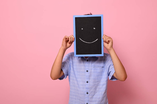 Unrecognizable school boy covers his face with a chalkboard with drawn smiling emoticons, expressing happiness. Isolated over pink background with copy space - Photo, image