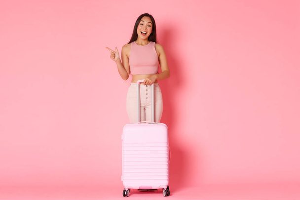 Reisen, Urlaub und Urlaubskonzept. Ganzkörperlange amüsierte schöne asiatische Mädchen in Sommerkleidung, Länder erkunden, den Sommer im Ausland genießen, nach links zeigen, mit Koffer stehen, rosa Hintergrund - Foto, Bild