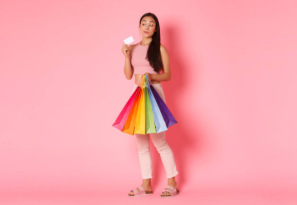 Full length portrait of thoughtful silly asian girl gonna spend all money on shopping, looking at credit card intrigued and holding bags with new clothes, standing over pink background - Photo, Image