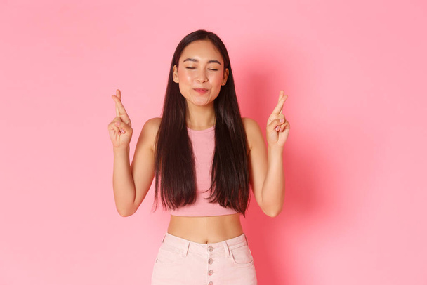 Lifestyle, beauty and women concept. Portrait of hopeful, happy asian girl in summer clothes making wish, cross fingers good luck smile and close eyes, praying over pink background - Photo, Image