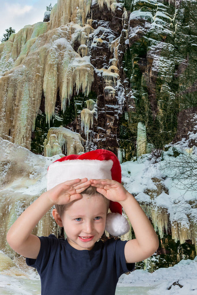 Multicolored ice streams on a steep cliff. Adorable little boy with blue eyes in a red Santa Claus hat is smiling. New Year in Finland. The concept of ecological and photo tourism - Foto, immagini