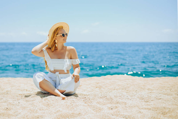Happy girl in white outfit sits on sand against the backdrop of the sea or ocean beach. Woman smiles and laughs, vacation and joy. Fashion model, beautiful jewelry, earrings, hat, sun glasses. - Foto, Imagem