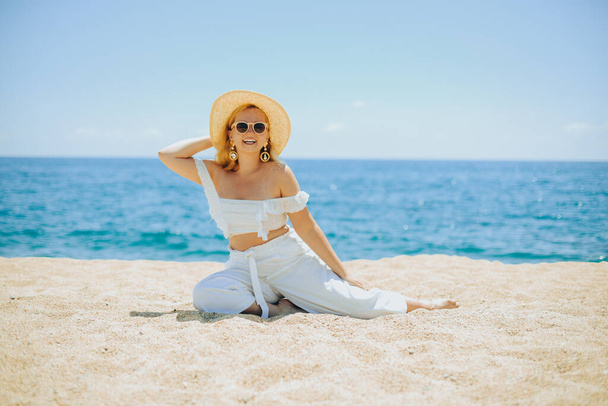Szczęśliwa dziewczyna w białym stroju siedzi na piasku na tle morza lub plaży oceanu. Kobieta uśmiecha się i śmieje, wakacje i radość. Modelka, piękna biżuteria, kolczyki, kapelusz, okulary przeciwsłoneczne. - Zdjęcie, obraz