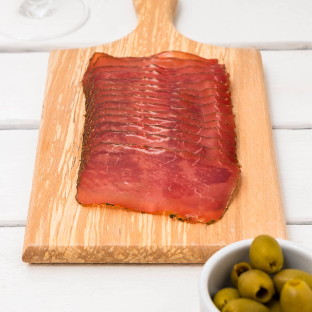 Tabla de cortar con salami seco en rodajas y aceitunas sobre mesa blanca - Foto, imagen