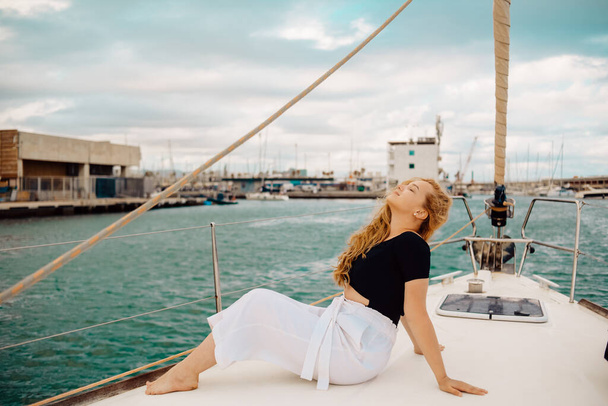 A beautiful long-haired girl sits on the bow of a barred yacht and enjoys life. The blonde is resting in the open sea or ocean. - Photo, Image