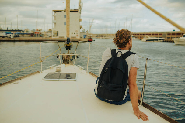 Guy utazó hátizsákkal ül egy vitorlás jachton, és élvezi a kilátást. Egy férfi egy jachton az óceánban, nyári napon. - Fotó, kép