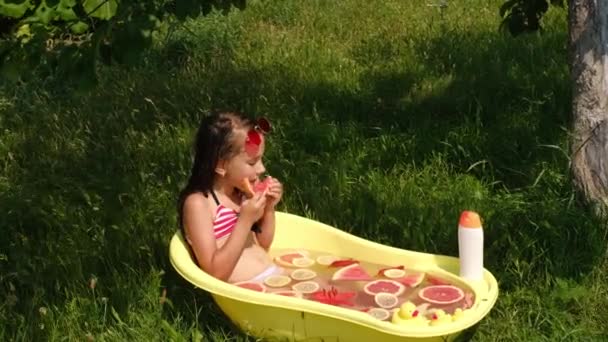 Dívka se koupe v přírodě na čerstvém vzduchu v zahradě. Koupel s plátky a kousky citrusových plodů a lilií. Užitečné přírodní oleje pro zdraví pokožky a těla.Dítě jí grapefruit - Záběry, video