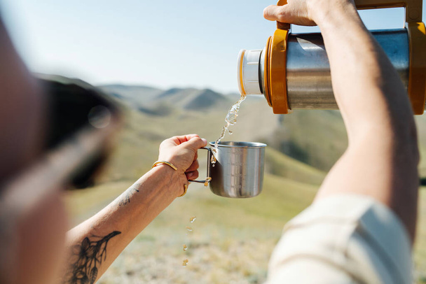Woman pouring tea from a thermos, he took on the hike. Over the shoulder view, blured head. Beautiful hills around - 写真・画像