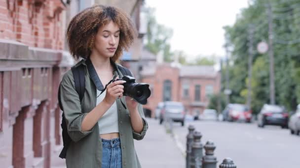 Positive young African American girl tourist visiting picturesque city street destination, using camera taking pictures on summer holiday, outdoors. Black biracial woman travel technology recreation - Footage, Video