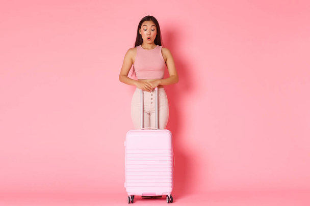 Matkailu, loma ja loma käsite. Täyspitkä huvittunut ja yllättynyt aasialainen kaunis tyttö kesävaatteissa näyttää hämmästynyt matkalaukku, tuijottaa alas, seisoo vaaleanpunainen tausta - Valokuva, kuva