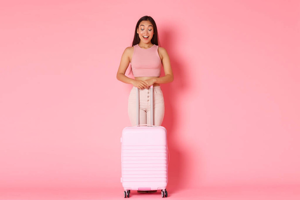 Путешествие, отпуск и концепция отпуска. Полная длина взволнованной и впечатленной азиатской привлекательной девушкой, смотрящей вниз с изумленной счастливой улыбкой, стоящей с чемоданом на розовом фоне - Фото, изображение