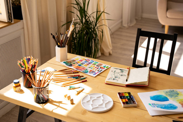 The artist's workplace with watercolor paints, gouache, brushes and sheets of paper on a wooden background. Copy space. Flat lay, top view. - 写真・画像