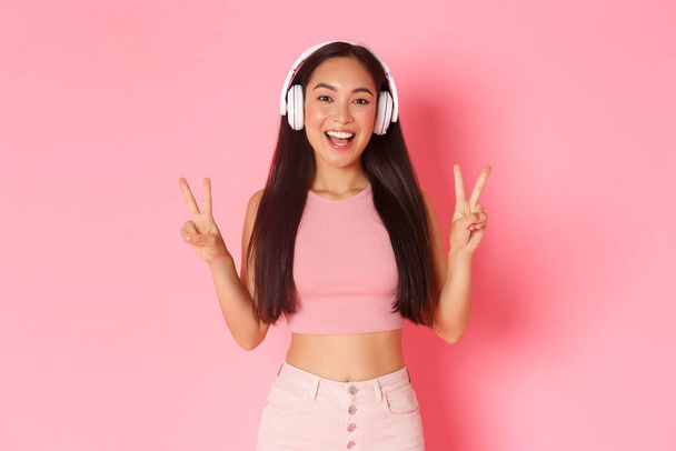 美しいとアップビートの肖像,ヘッドフォンで音楽を聞いて笑顔アジアの女の子,平和カワイイジェスチャーを示す,正のビブを送信,ピンクの背景の上に立って - 写真・画像