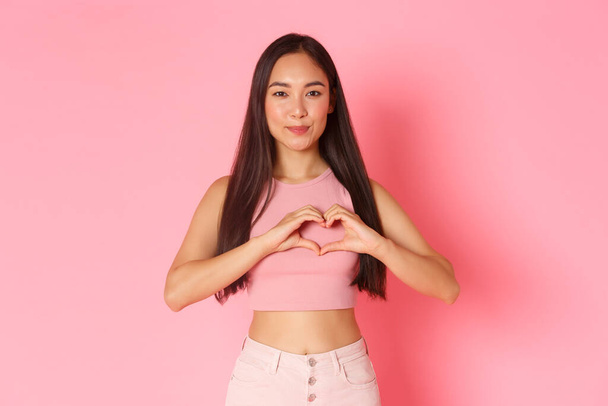 Lifestyle, beauty and women concept. Portrait of attractive smiling, happy asian girl confess in sympathy, like someone, showing heart gesture as feeling love, pink background - Foto, afbeelding