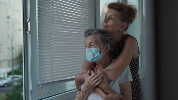Einsame Seniorin 90 Jahre alt in medizinischer Maske und ihre reife Tochter in Umarmung, die aus dem Fenster im Pflegeheim blickt. Erwachsene Tochter besucht ältere Mutter liebt sie sehr und unterstützt sie. - Foto, Bild