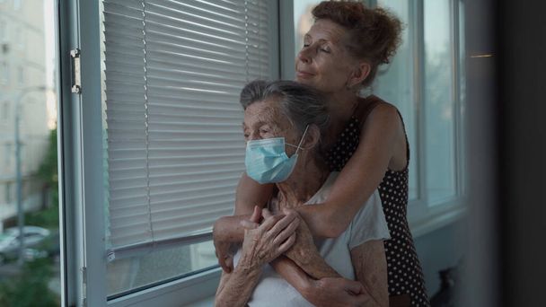 Volwassen dochter omarmt met liefdevolle senior moeder 90 jaar in masker voor het raam, familieleden van verschillende generaties, vrouwen in omhelzing, dromen van geluk, tijd samen doorbrengen. - Foto, afbeelding