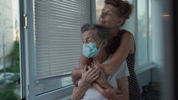 Einsame Seniorin 90 Jahre alt in medizinischer Maske und ihre reife Tochter in Umarmung, die aus dem Fenster im Pflegeheim blickt. Erwachsene Tochter besucht ältere Mutter liebt sie sehr und unterstützt sie. - Foto, Bild