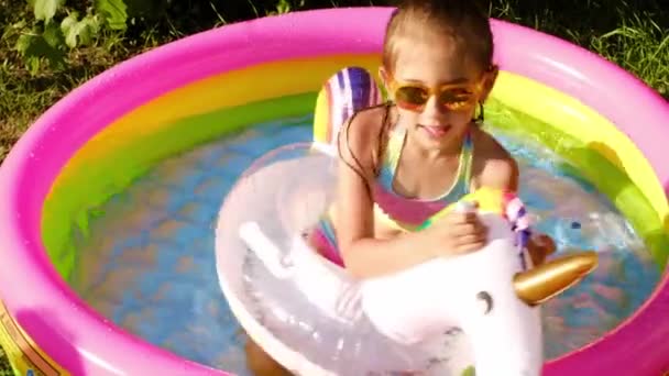 Uma menina em óculos de sol frolics em uma pequena piscina inflável com um círculo na forma de um unicórnio no jardim. Brincadeiras infantis em um dia quente de verão - Filmagem, Vídeo
