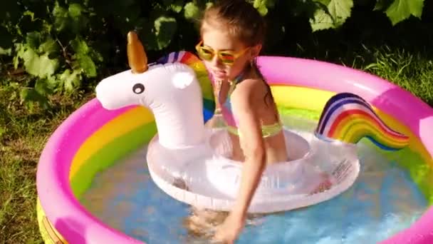 Egy napszemüveges gyerek úszik egy unikornis alakú mentőövvel egy kis felfújható medencében. Egy lány tiszta vízben fröcsköl egy forró nyári estén. - Felvétel, videó