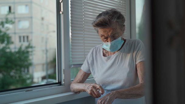 Stará žena 90 let s obličejovou maskou používá alkohol sprej chránit covid 19 stojí u okna v pečovatelském domě, zůstat doma, zdravotní koncepce, nový normální. Hygiena rukou při izolaci koronaviru. - Fotografie, Obrázek
