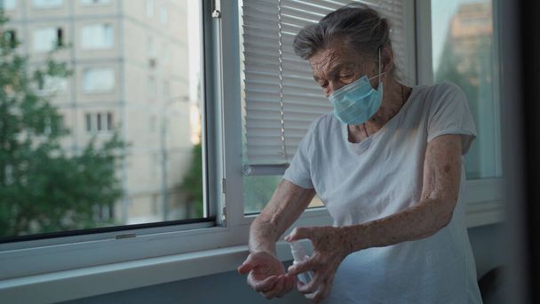 90歳の女性の顔のマスクを使用してアルコールスプレーを身に着けている家庭で窓のそばに立って、家庭での滞在、医療の概念は、新しい正常なcovid 19を保護します。コロナウイルスの分離中に手の衛生. - 写真・画像