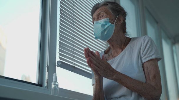 Elderly woman in a medical mask washing hands with alcohol gel or antibacterial soap sanitizer standing by the window in a nursing home. Hygiene during the COVID-19 coronavirus pandemic. - Фото, зображення