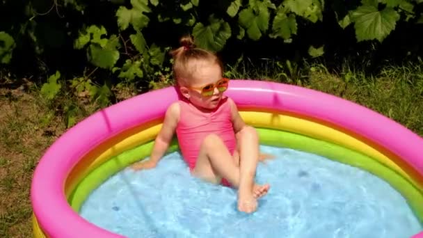 Una bambina schizza in acqua in una piccola piscina gonfiabile in giardino. Una calda giornata di sole con attività acquatiche. Mini piscina portatile per bambini - Filmati, video