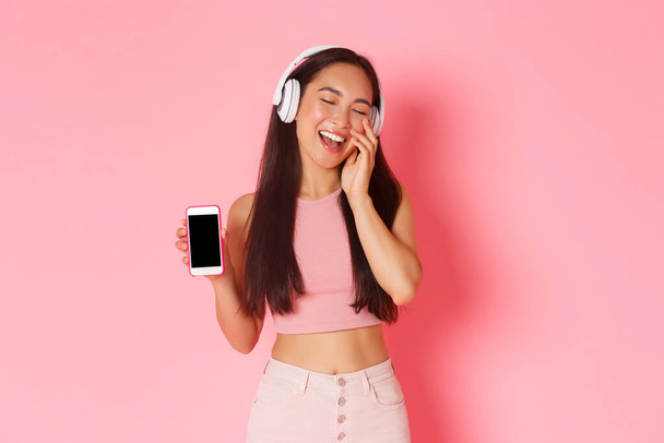 Technologie, Kommunikation und Online-Lifestyle-Konzept. Porträt einer wunderschönen, unbeschwerten asiatischen Frau, die die Augen schließt und süß lächelt, während sie Musik über Kopfhörer hört und den Handybildschirm zeigt - Foto, Bild