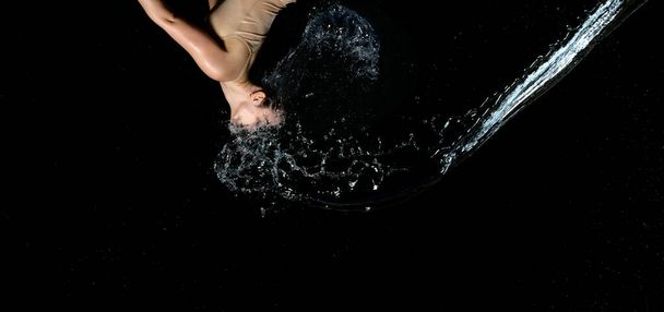 beautiful woman of Caucasian appearance with black hair in drops of water on a black background. A jet of water flies in the face - Zdjęcie, obraz