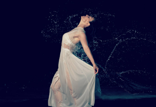 красивая женщина кавказской внешности с черными волосами танцует в каплях воды на черном фоне. Женщина одета в белое шифонское платье - Фото, изображение