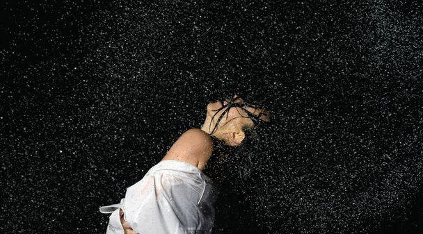 beautiful woman of Caucasian appearance with black hair dances in drops of water on a black background. Woman wearing white shirt - Фото, изображение