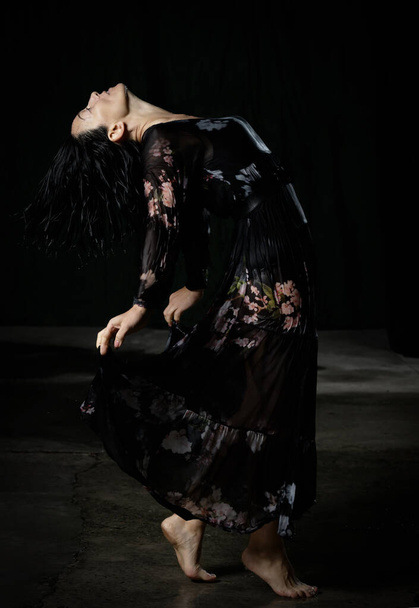 νεαρή γυναίκα με μαύρα μαλλιά χορεύει σε μαύρο βρεγμένο μακρύ φόρεμα σιφόν σε σκούρο φόντο. Η γυναίκα είναι λυγισμένη πίσω. - Φωτογραφία, εικόνα