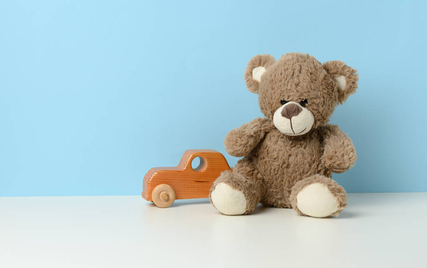 Niedlicher brauner Teddybär sitzt auf einem weißen Tisch und einem hölzernen Kinderspielzeugauto, blauer Hintergrund - Foto, Bild