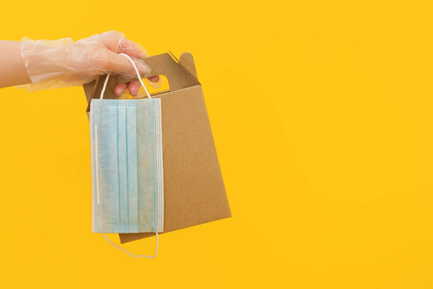 a hand in a disposable rubber glove holds a cardboard box and a face mask on a yellow background, contactless delivery concept, blank packaging for your design - Foto, Imagem