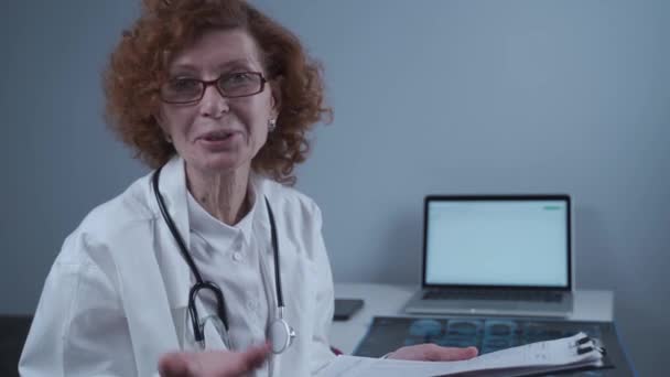 Senior woman doctor speaks looking into camera holding clipboard in hands sitting in medical office near computer. Elderly female general practitioner advises patient while reviewing medical tests - Séquence, vidéo
