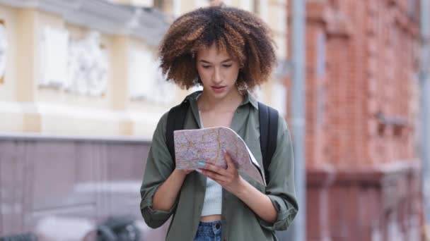 Θετική νεαρή Αφρο-Αμερικανίδα πεζοπόρος με σακίδιο που κρατάει ταξιδιωτικό χάρτη. Ταξιδεύοντας σγουρά μαλλιά μαύρο κορίτσι φοιτητής έλεγχο κατεύθυνση στο χάρτη χαρτί εξερεύνηση κατοπτεύσεις στην Ευρώπη - Πλάνα, βίντεο