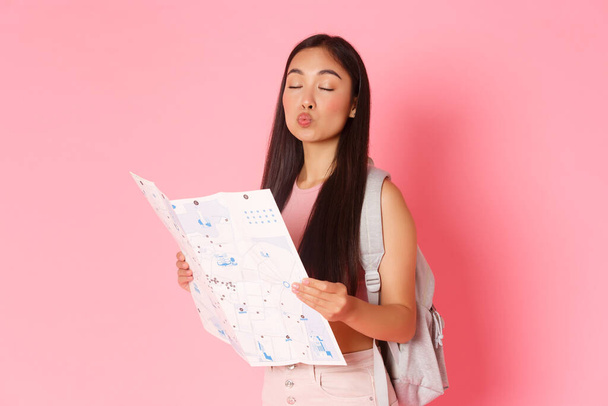 Viajar, estilo de vida y concepto turístico. Retrato de chica asiática tonta turista con mochila y mapa, ojos cerrados y haciendo pucheros besándose la cara, buscando ruta de vacaciones, fondo rosa - Foto, imagen