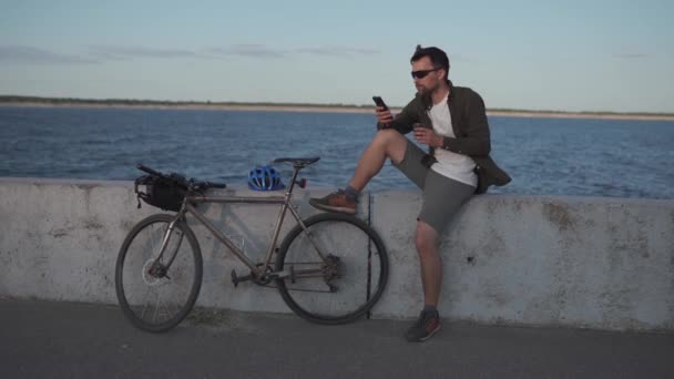 自転車での旅行者は、海へのスマートフォンのビューとチェックロードマップを楽しむために停止し、旅行のサイクルタイムでコーヒーを持っています。男性サイクリストはスマートフォンとドリンクを使って堤防でコーヒーを飲みます - 映像、動画