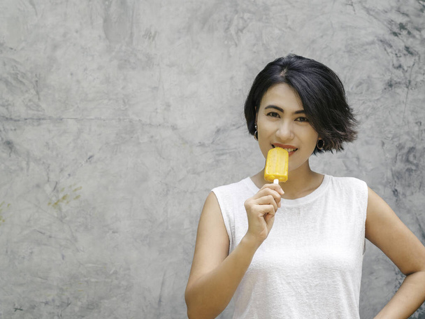 Uśmiechnięta Azjatka krótkie włosy w luźnej koszuli bez rękawów jedząca żółtego loda na szarym betonowym tle latem. Kobiety jedzące lody z przestrzenią do kopiowania. - Zdjęcie, obraz