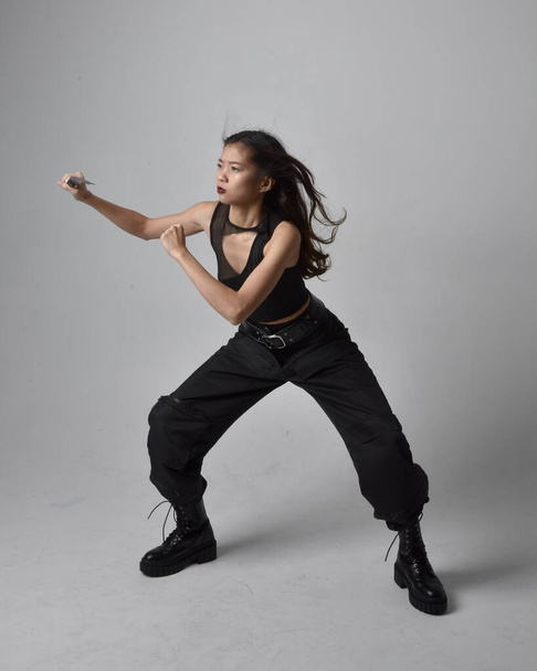 Full length portrait of pretty young asian girl wearing black tank top, utilitarian  pants and leather boots. Standing pose holding a knife,  isolated against a  studio background. - Zdjęcie, obraz