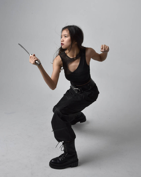 Full length portrait of pretty young asian girl wearing black tank top, utilitarian  pants and leather boots. Standing pose holding a knife,  isolated against a  studio background. - Фото, зображення