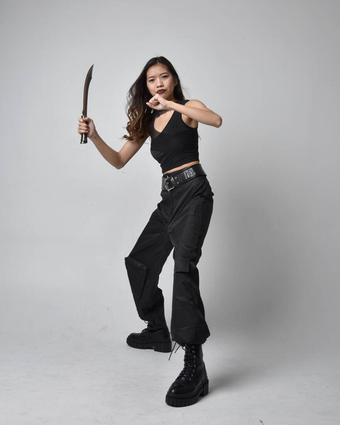 Full length portret van mooie jonge aziatische meisje dragen zwarte tank top, utilitaire broek en lederen laarzen. Staande houding met een mes, geïsoleerd tegen een studio achtergrond. - Foto, afbeelding