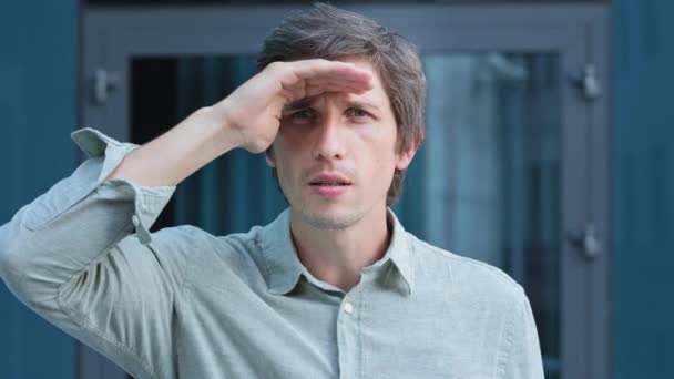 Headshot Portrait of man looking suspicious, gazes intently, peers into distance, covers face from sun with his palm, expects meeting. Doubtful, frustrated person mistrustful, screwing up his eyes - Footage, Video