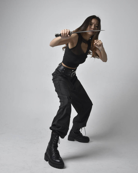 Full length portrait of pretty young asian girl wearing black tank top, utilitarian  pants and leather boots. Standing pose holding a knife,  isolated against a  studio background. - Fotó, kép
